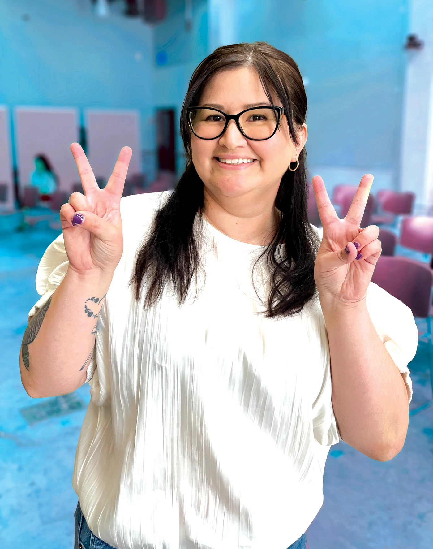 Maria Martinez smiles and holds up peace signs in front of a blurred CSUN classroom or campus interior.