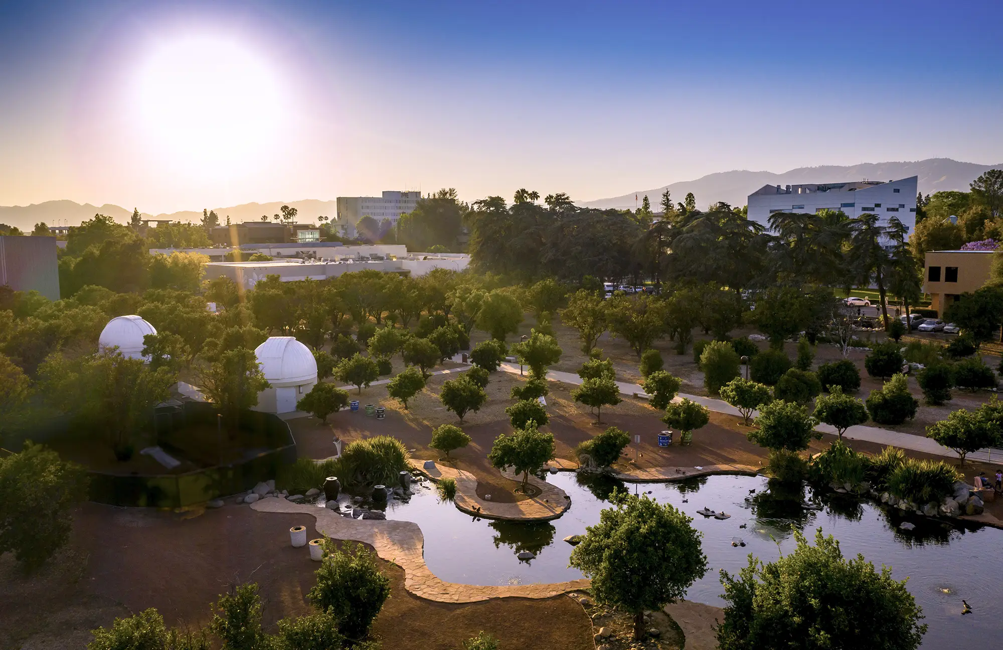 Aerial view of the CSUN campus at sunset, showing a pond, white observatory domes, and trees with mountains in the background.