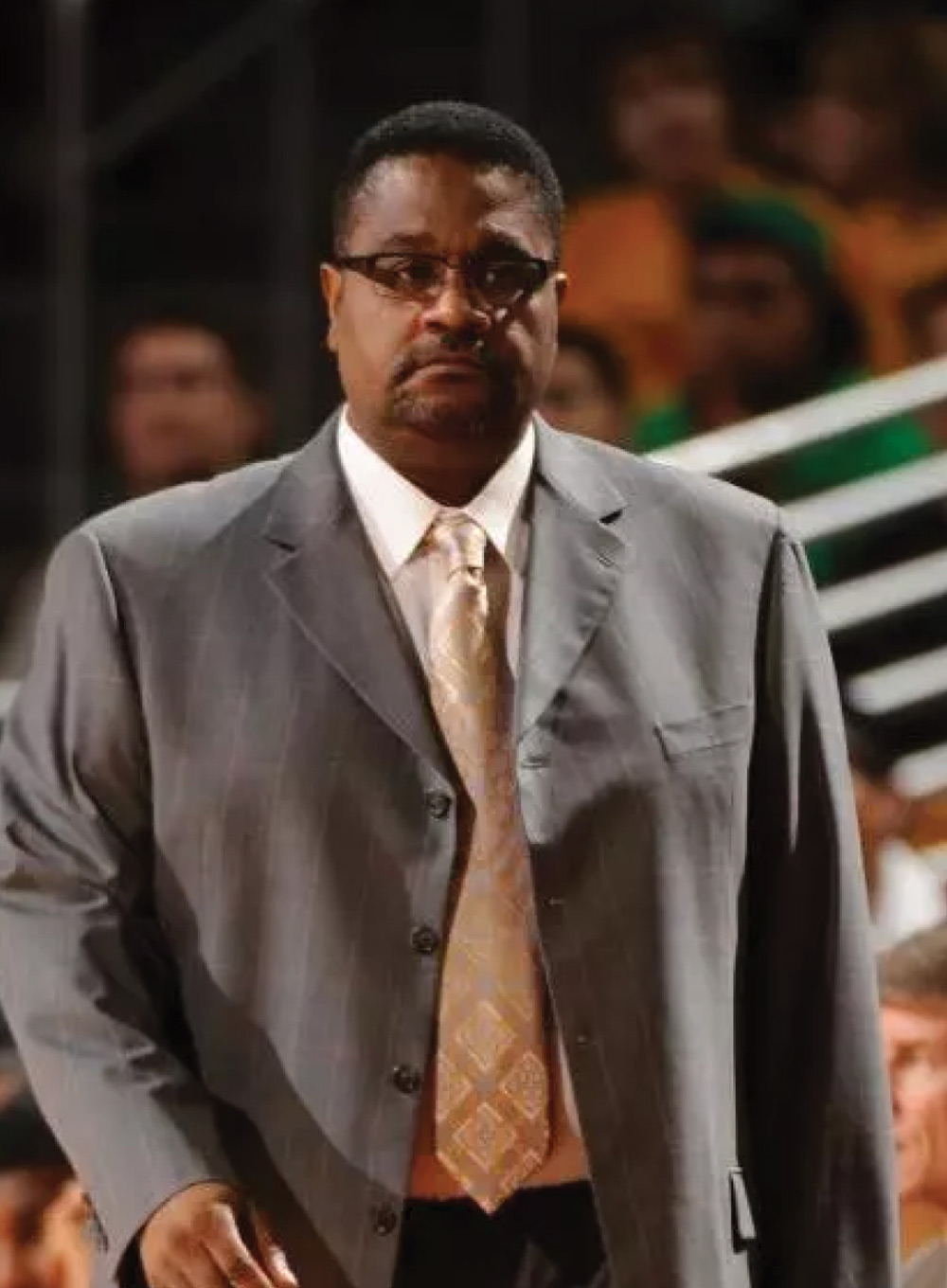 Bobby Braswell in a grey suit, patterned tie, and glasses standing indoors looking focused.