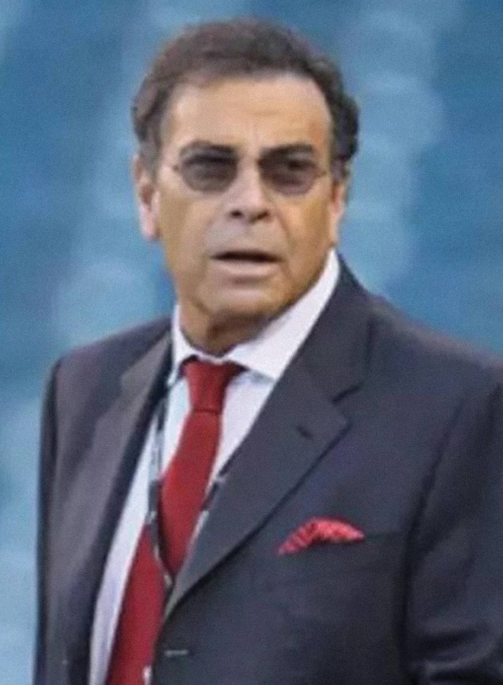 Dennis Gilbert wearing a dark suit jacket, red tie, and tinted glasses.