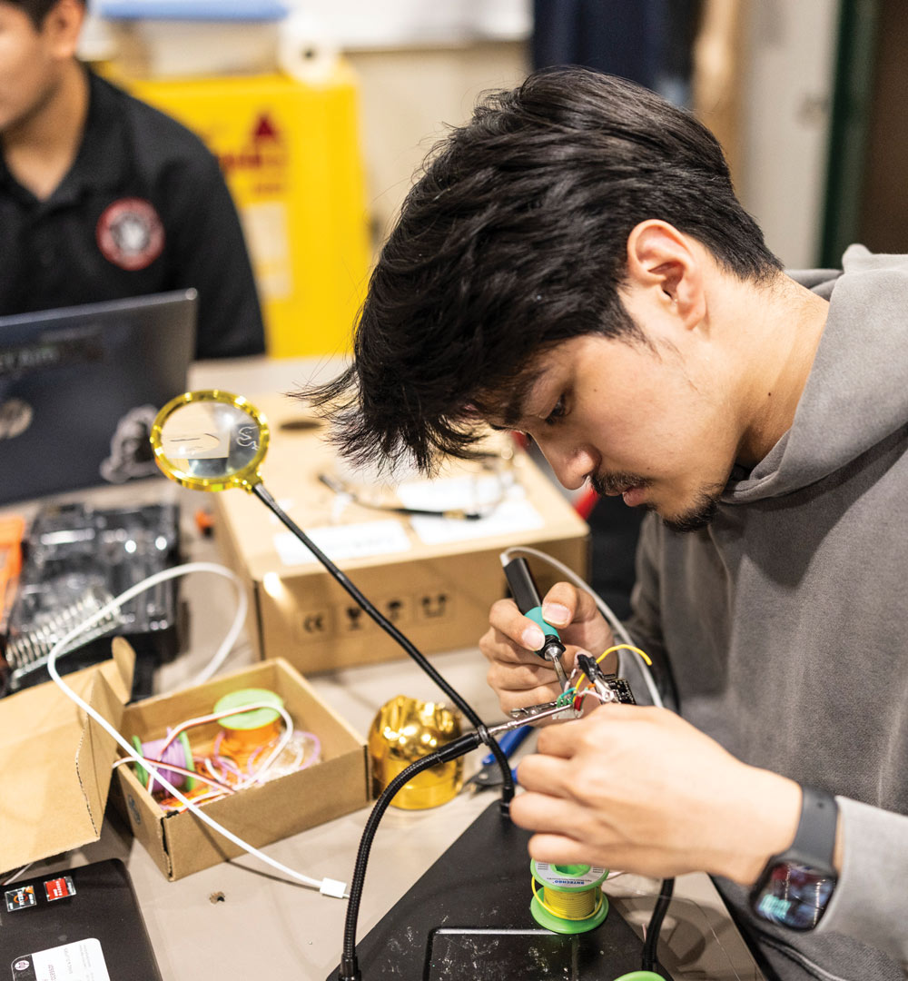 An engineering student carefully soldering small electronic components on a workbench in a lab.
