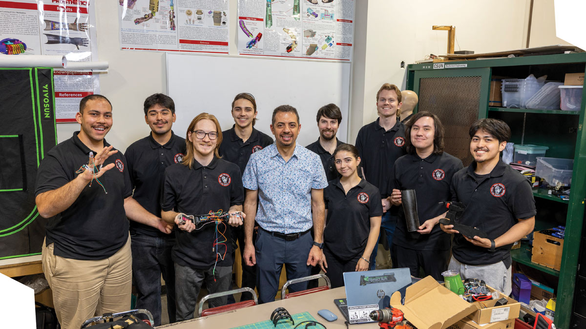 A professor and group of engineering students posing together in a workshop holding prosthetic hand prototypes.