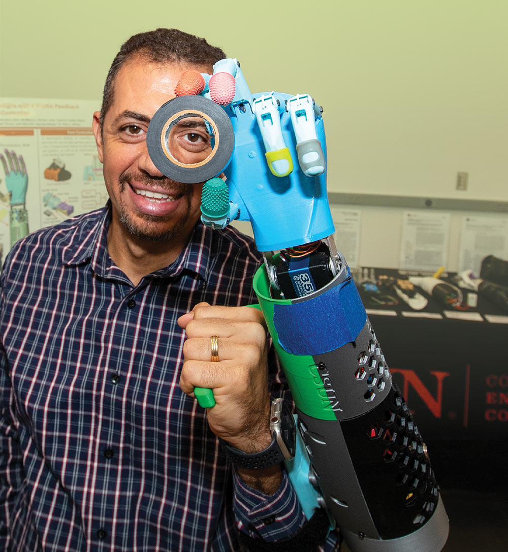 Peter Bishay smiling behind a colorful 3D-printed prosthetic arm that he is holding up in a laboratory setting.