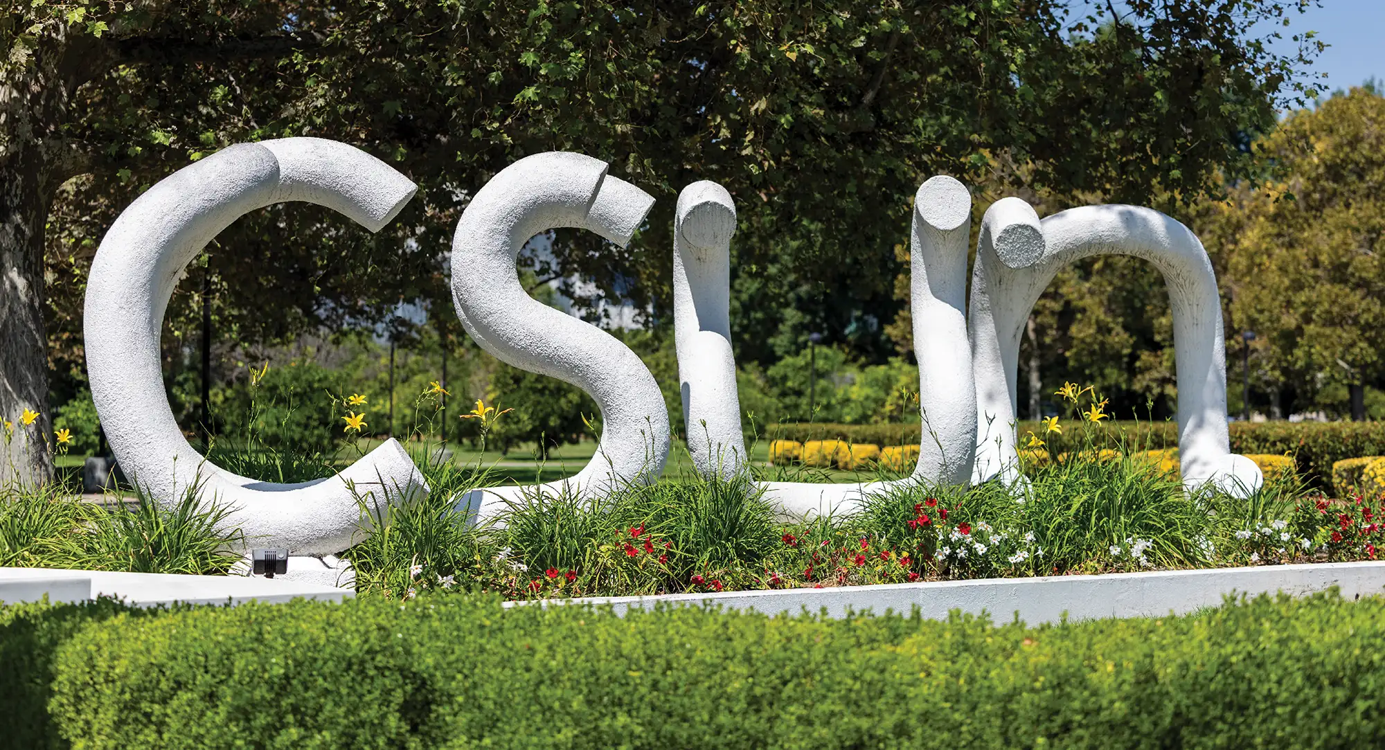 Large white sculpture spelling "CSUN" surrounded by green plants and flowers on a campus lawn.