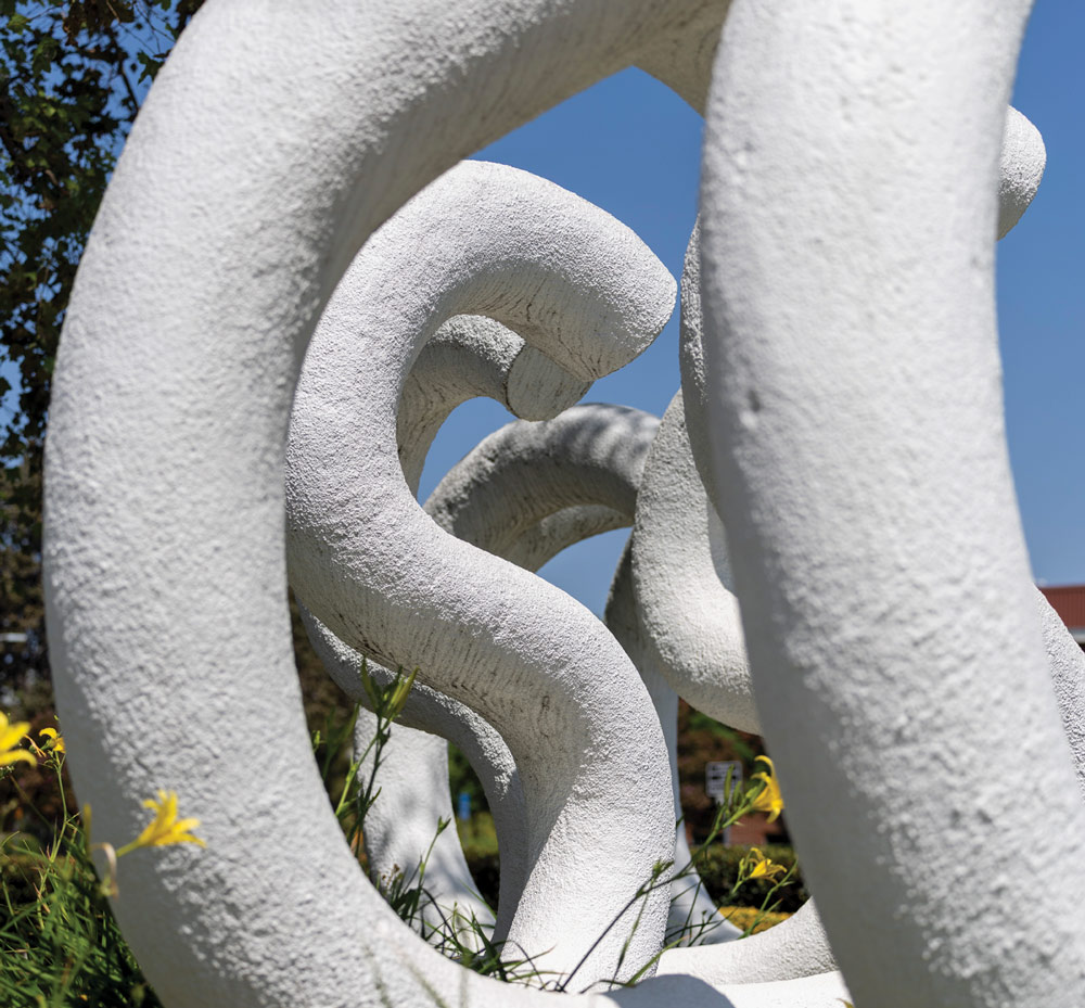 Close-up perspective of the white CSUN letter sculpture with yellow flowers in the foreground under a blue sky.