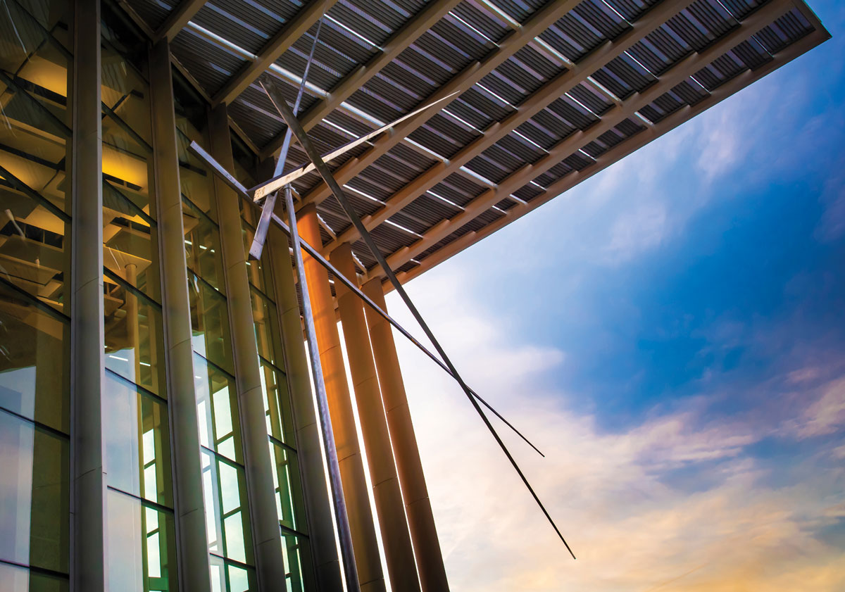 Modern architectural facade of a glass building with metallic overhangs and columns against a colorful evening sky.