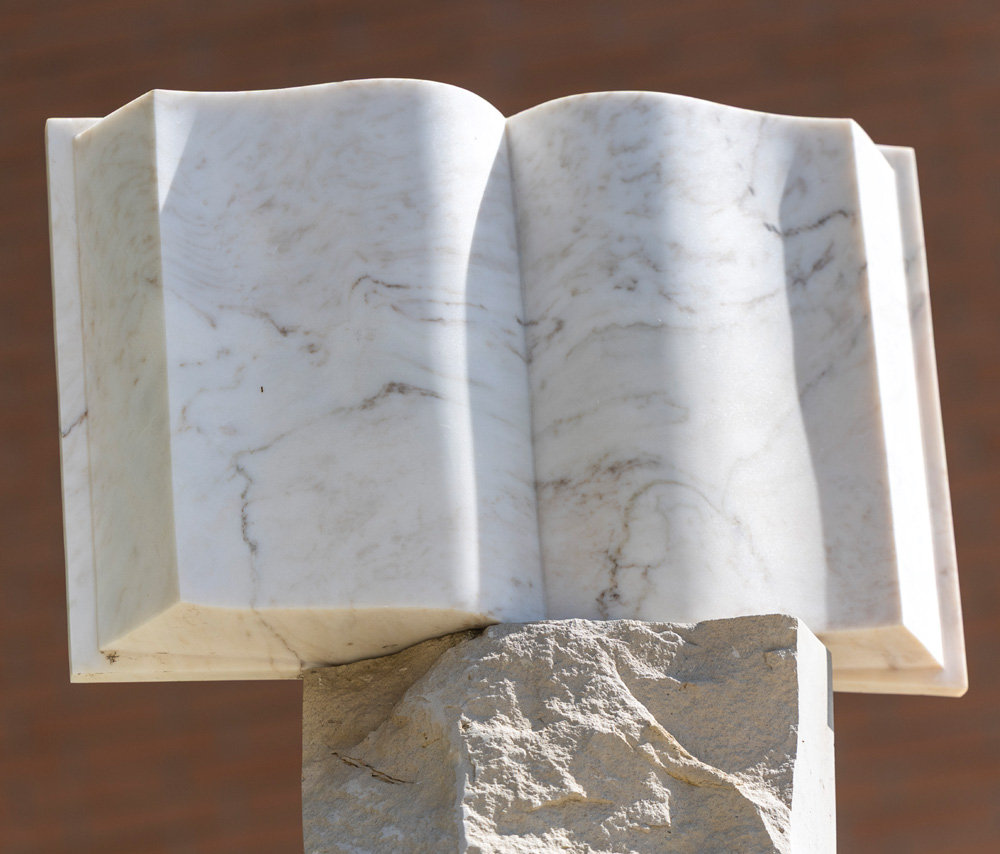 Close-up view of a marble sculpture depicting an open book resting on a stone pedestal.