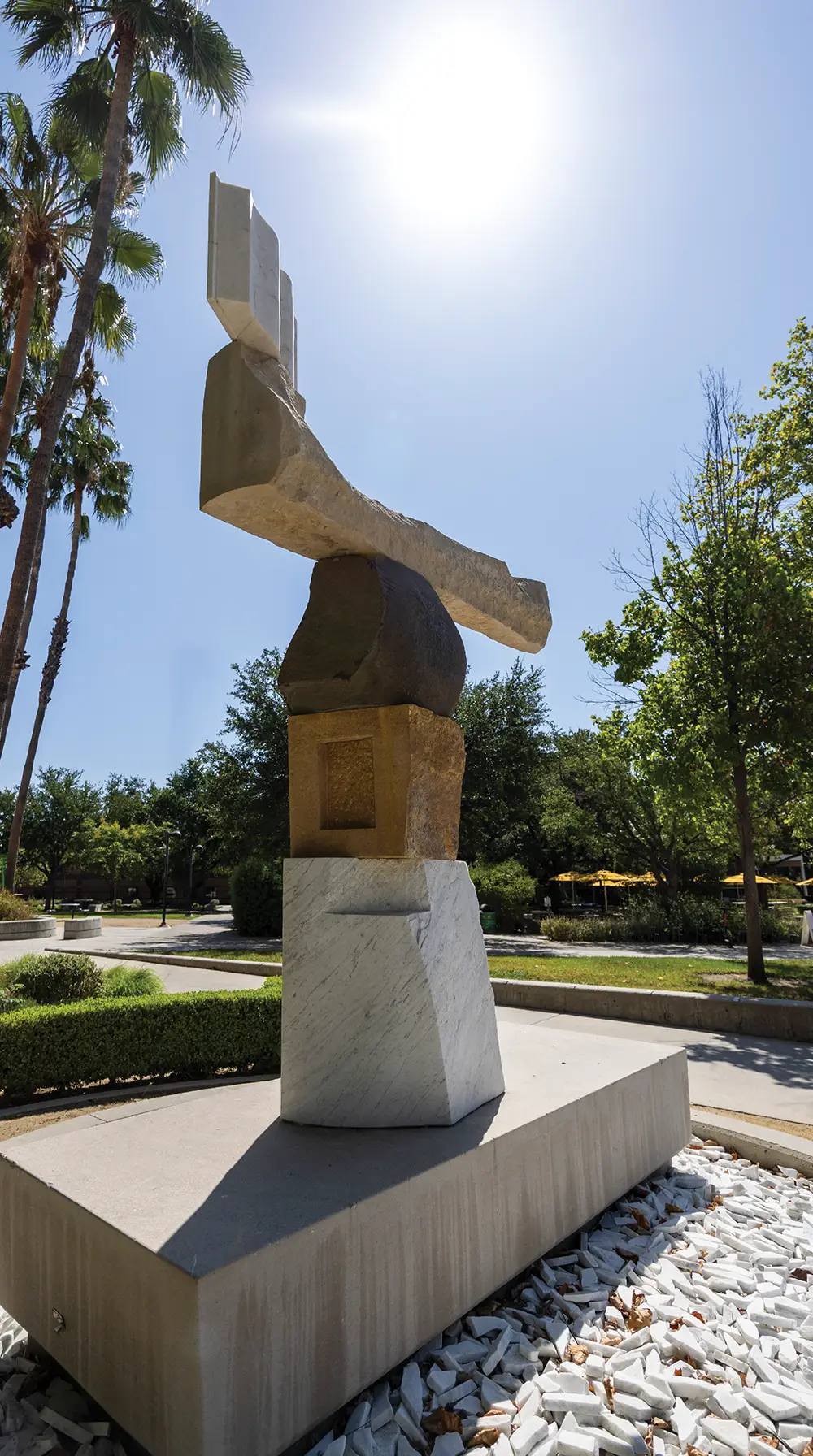 A tall, stacked stone sculpture topped with an open book design set outdoors on a campus plaza.