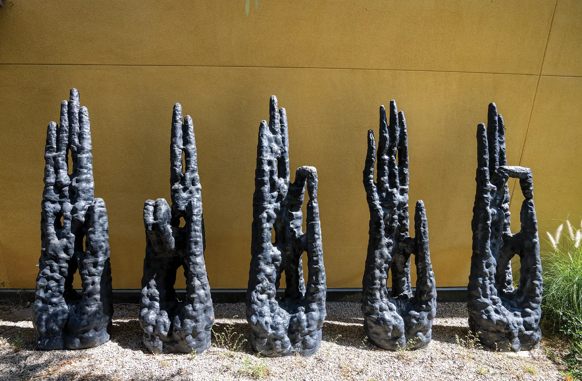 Five tall, dark, textured pillar sculptures standing in a line against a yellow wall outdoors.