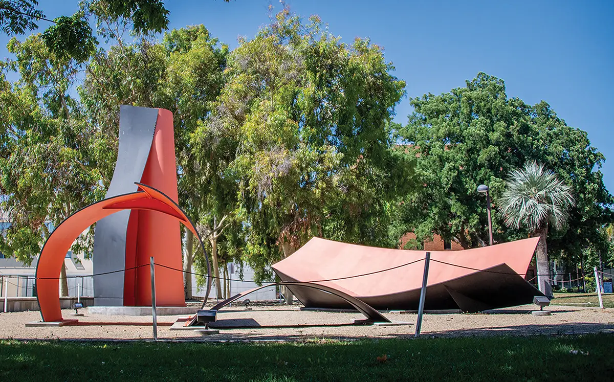 Large, orange-red metal abstract sculptures displayed outdoors in a grassy park setting with trees.