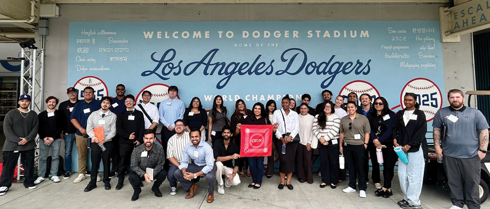 A large group of students and alumni pose in front of a "Welcome to Dodger Stadium" sign while holding a red CSUN flag.