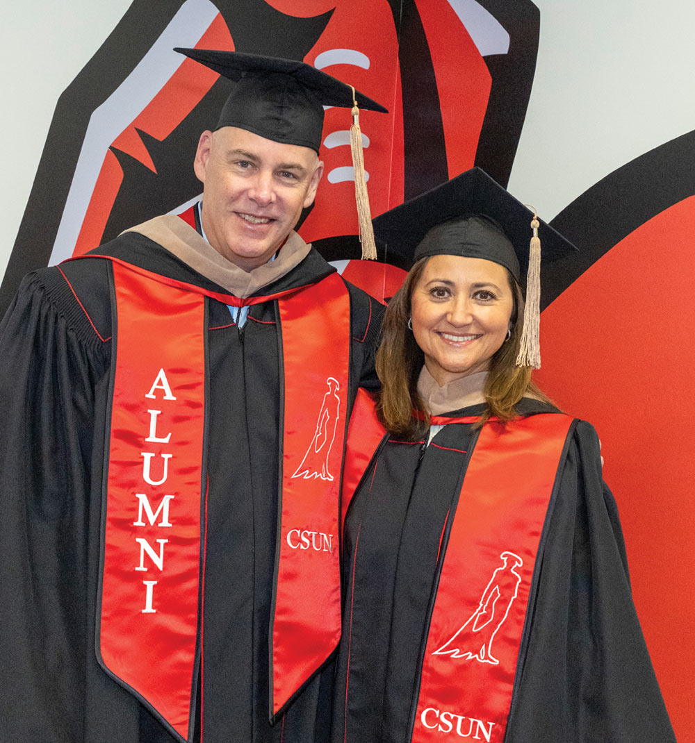 Elsa Burton and Jeff Burton wearing black graduation robes and red CSUN Alumni stoles smile together at a ceremony.