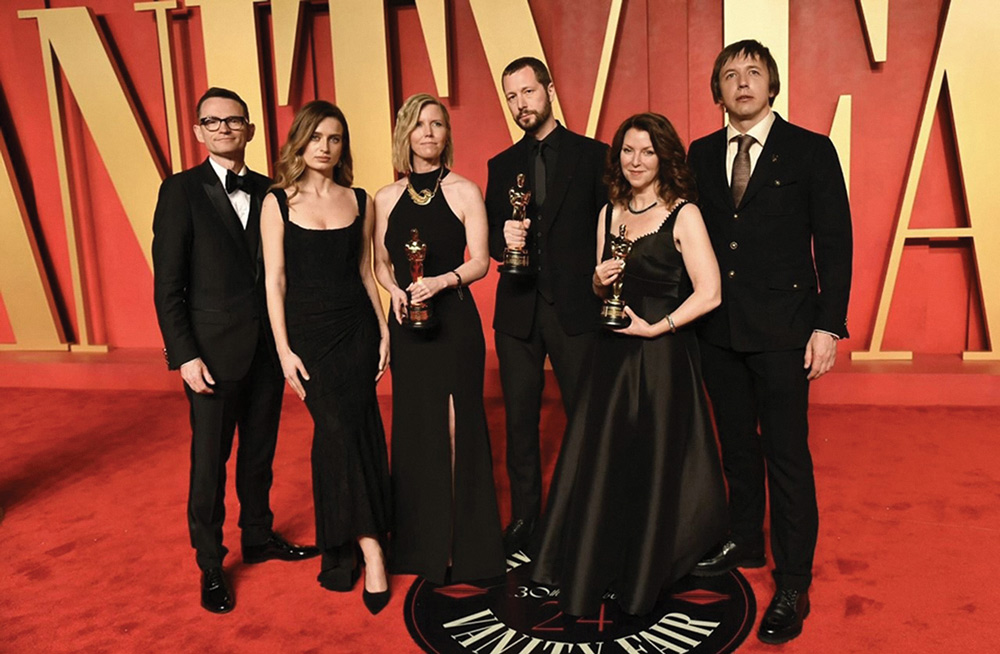 Michelle Mizner and five other people in formal black attire, three holding Oscar statues, pose together on a red carpet at a Vanity Fair event.