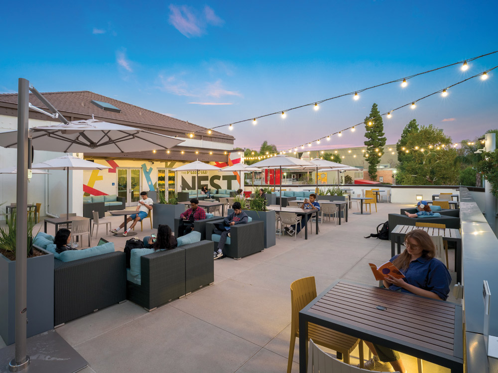 Students relax at outdoor lounge tables on a lit patio called "The Nest" under string lights at dusk.