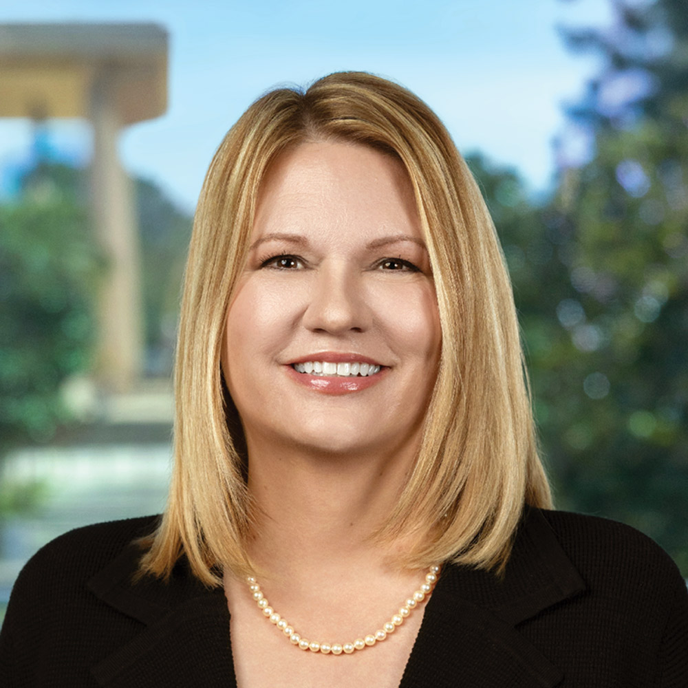 Erika D. Beck with blonde, shoulder-length hair wearing a pearl necklace and black jacket against a blurred outdoor backdrop.