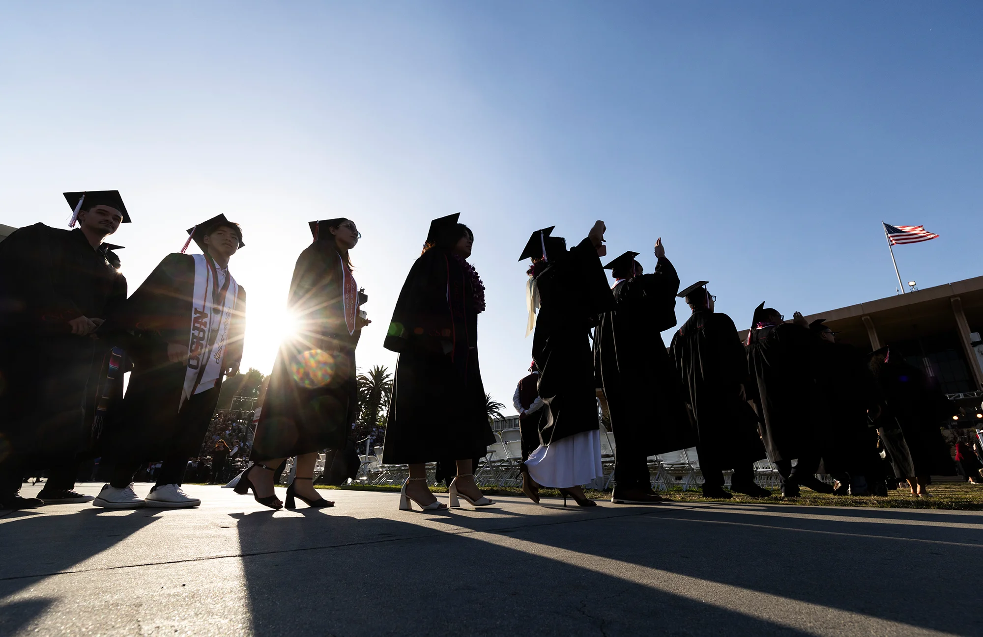 Graduates in caps and gowns walk in a line outdoors, silhouetted against bright sunlight with an American flag flying nearby.