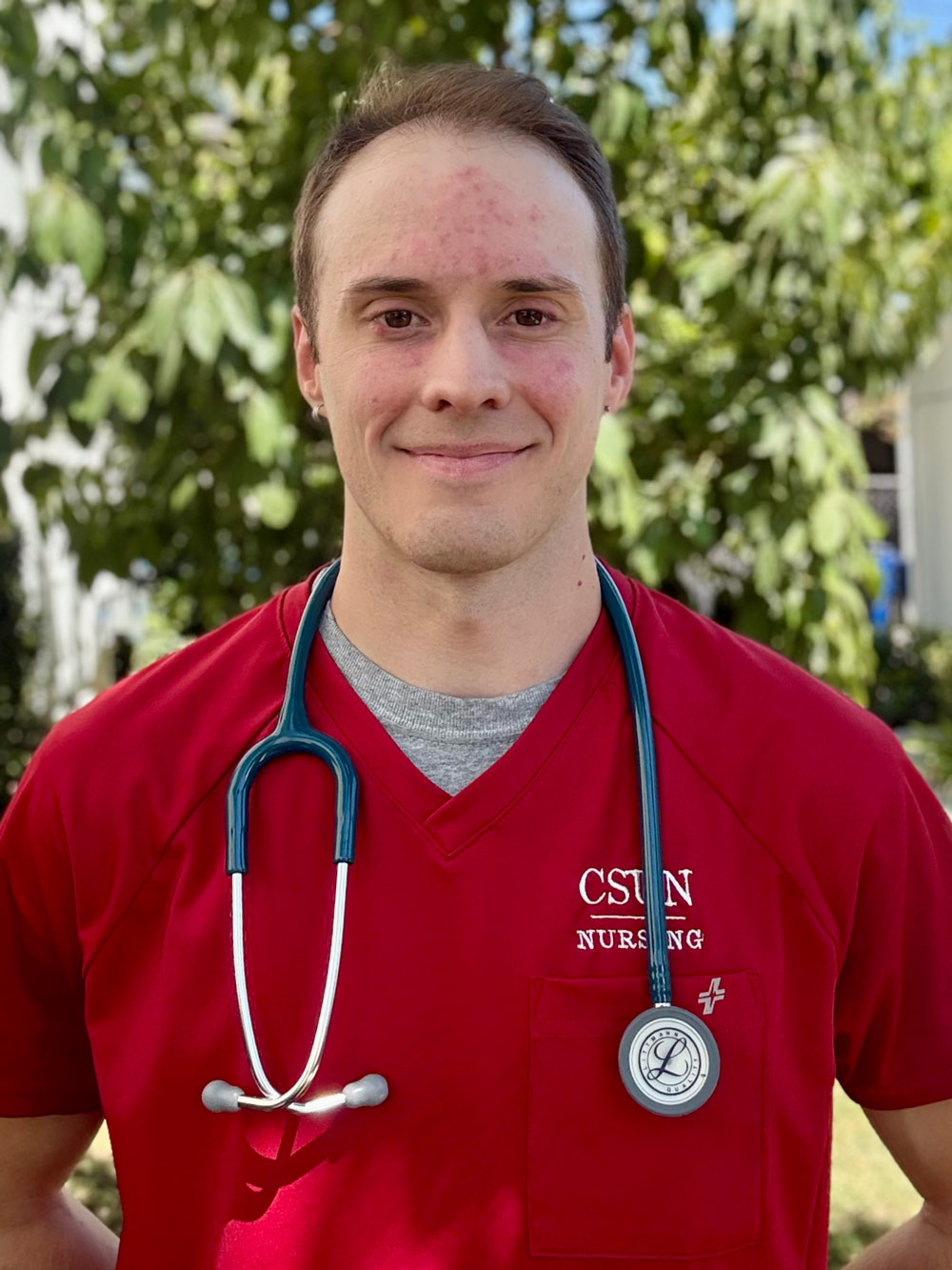 Daniel Rietz wearing red CSUN Nursing scrubs with a stethoscope around his neck smiles against a blurred background.