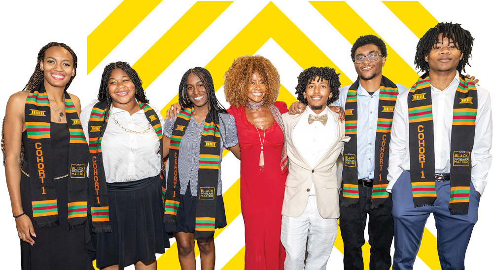 A group of six members of the Black Scholars Matter program wearing graduation stoles posing together indoors with an instructor in red.