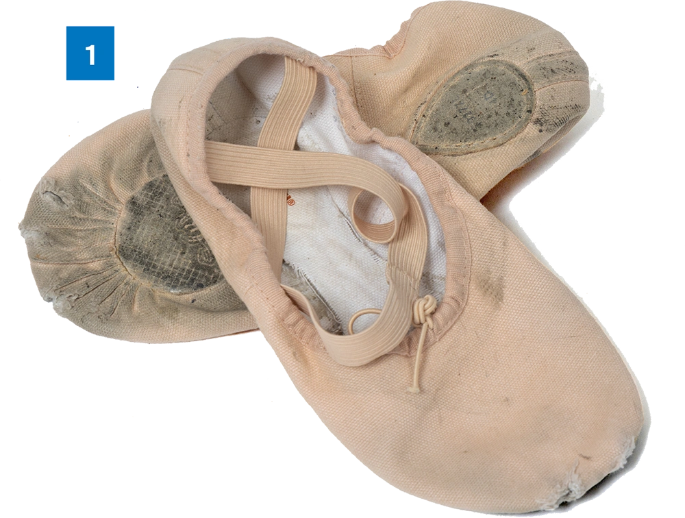 A pair of worn pink ballet shoes with crossed elastic straps laid on a white background, labeled with number 1.