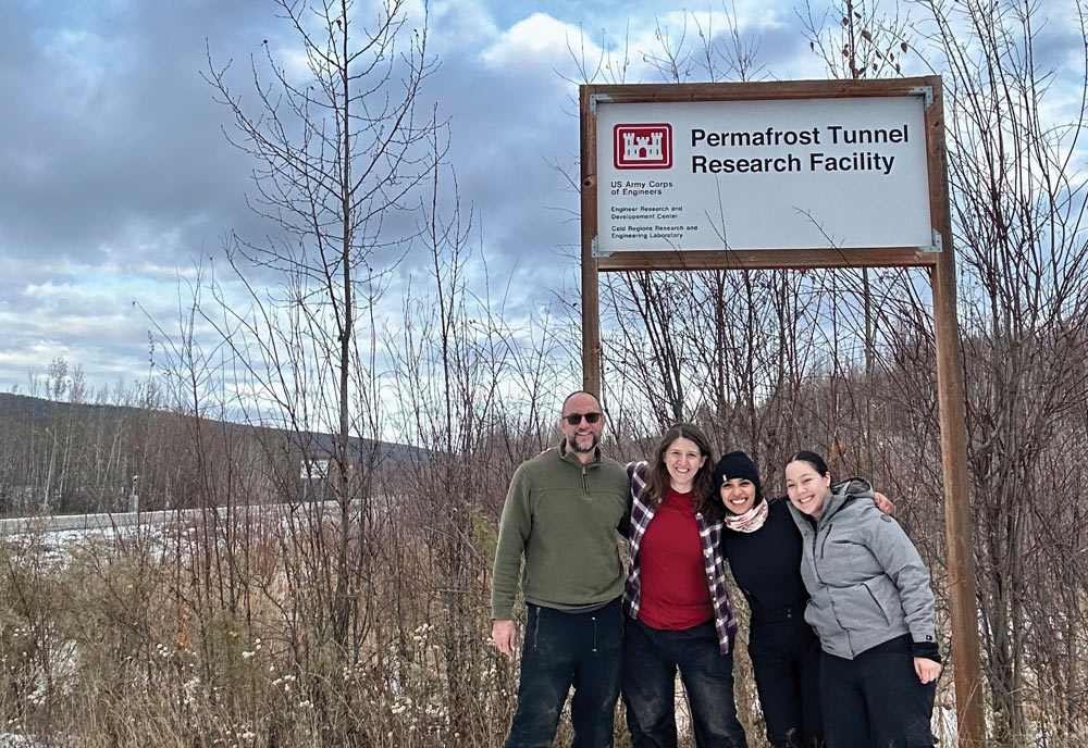 Four researchers pose together in winter gear in front of a Permafrost Tunnel Research Facility sign outdoors.