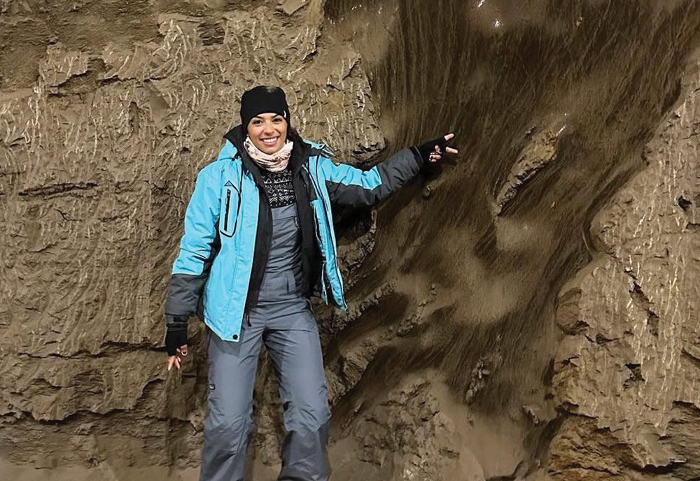 A woman in winter apparel smiles inside a permafrost tunnel, gesturing toward an icy, textured dirt wall.