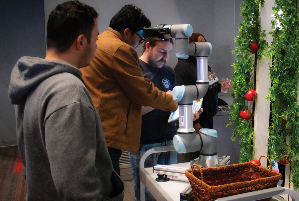 Students gather around a robotic arm operating next to a vine wall with red apples and a basket.
